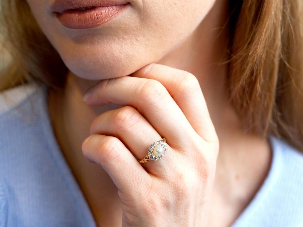An 18ct yellow gold cluster ring featuring a central oval natural pearl surrounded by a halo of old-cut diamonds with scroll-carved shoulders. View III