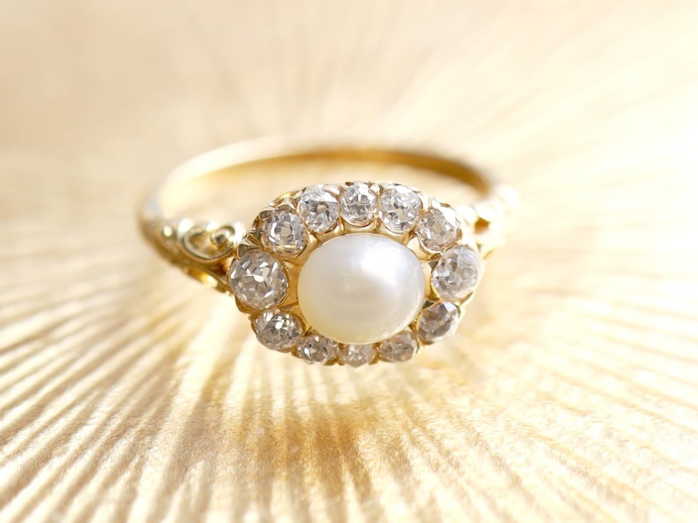An 18ct yellow gold cluster ring featuring a central oval natural pearl surrounded by a halo of old-cut diamonds with scroll-carved shoulders. View V