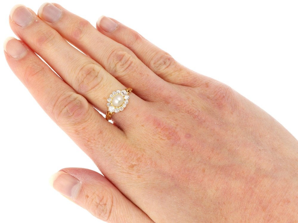 An 18ct yellow gold cluster ring featuring a central oval natural pearl surrounded by a halo of old-cut diamonds with scroll-carved shoulders. View X