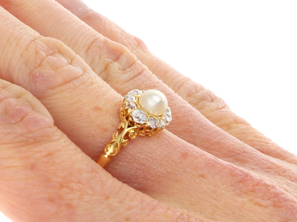 An 18ct yellow gold cluster ring featuring a central oval natural pearl surrounded by a halo of old-cut diamonds with scroll-carved shoulders. View XI
