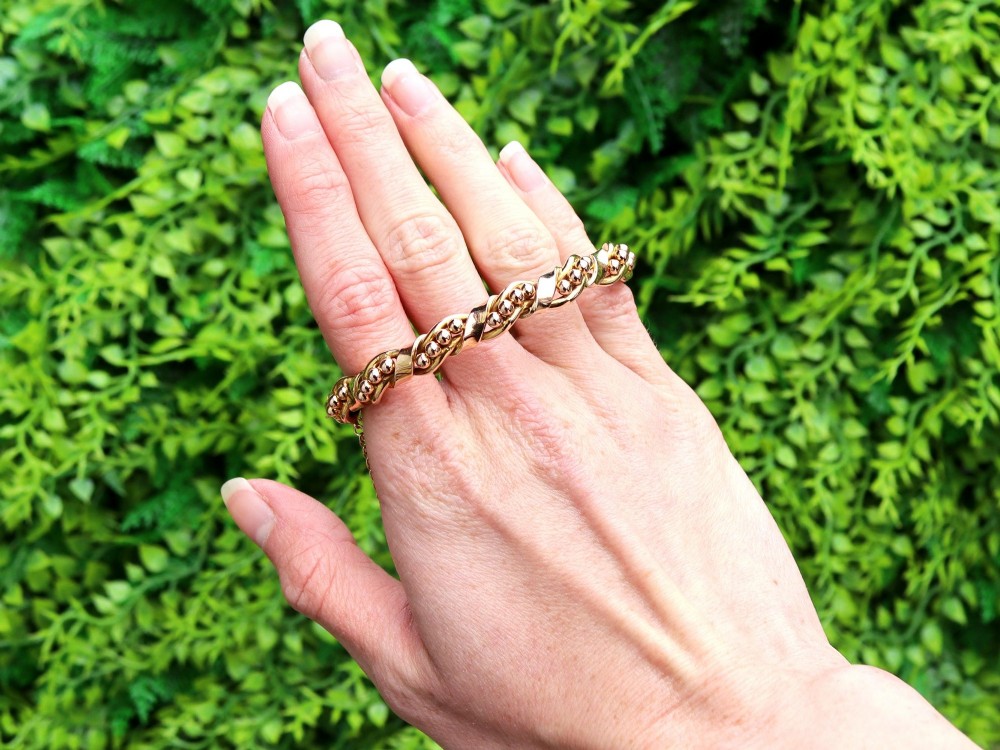 An antique Edwardian 15ct gold hinged bangle featuring a twisted ribbon design intertwined around a row of polished gold beads, fitted with a safety chain. View II