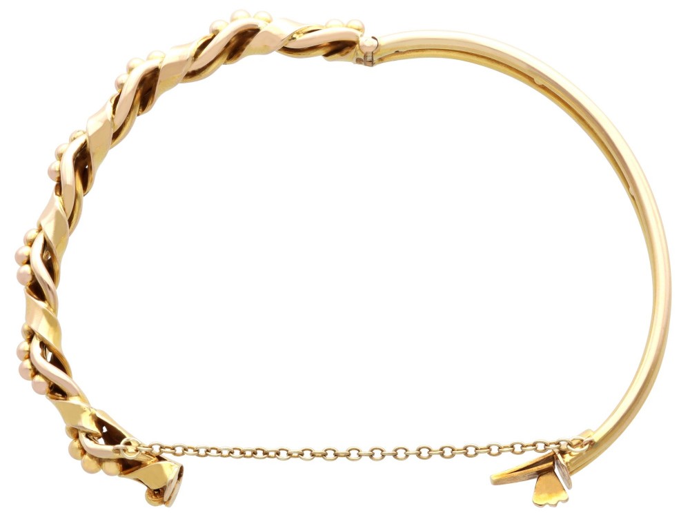 An antique Edwardian 15ct gold hinged bangle featuring a twisted ribbon design intertwined around a row of polished gold beads, fitted with a safety chain. View X