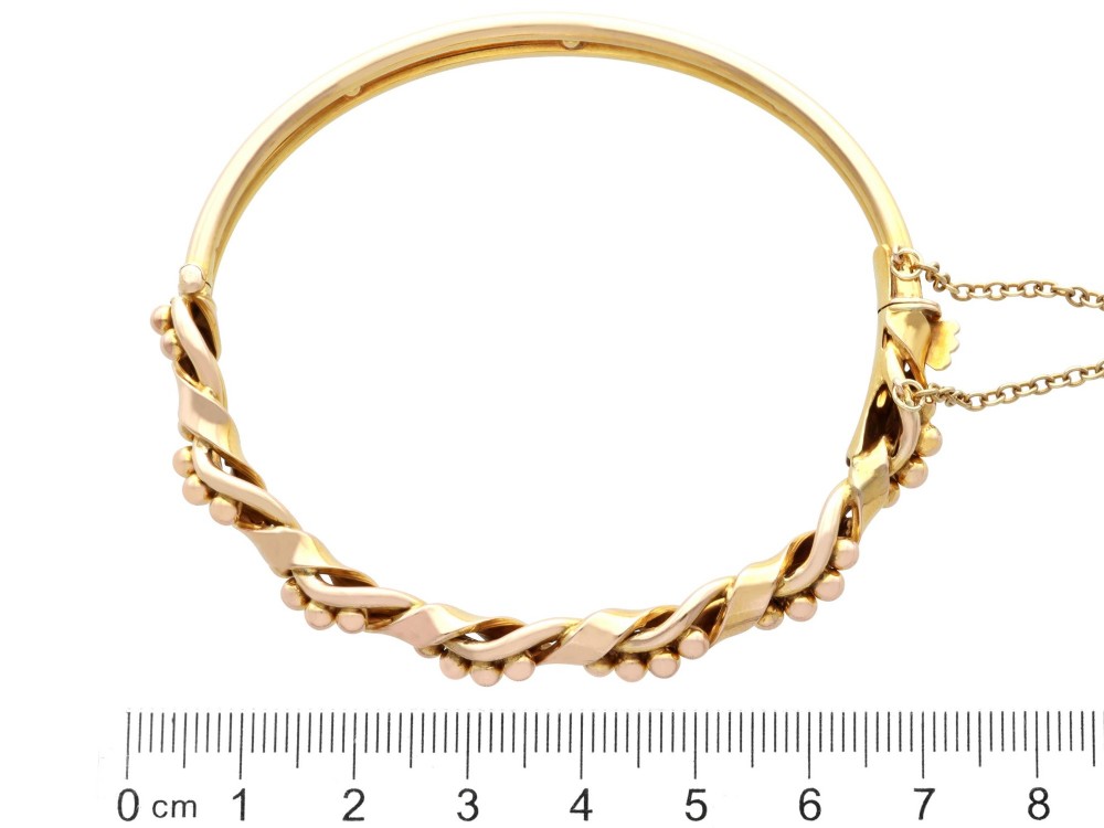 An antique Edwardian 15ct gold hinged bangle featuring a twisted ribbon design intertwined around a row of polished gold beads, fitted with a safety chain. View XI