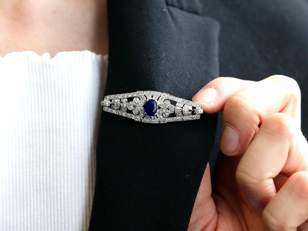 An Art Deco 9ct white gold brooch featuring a central oval blue sapphire framed by geometric openwork set with round diamonds. View IV