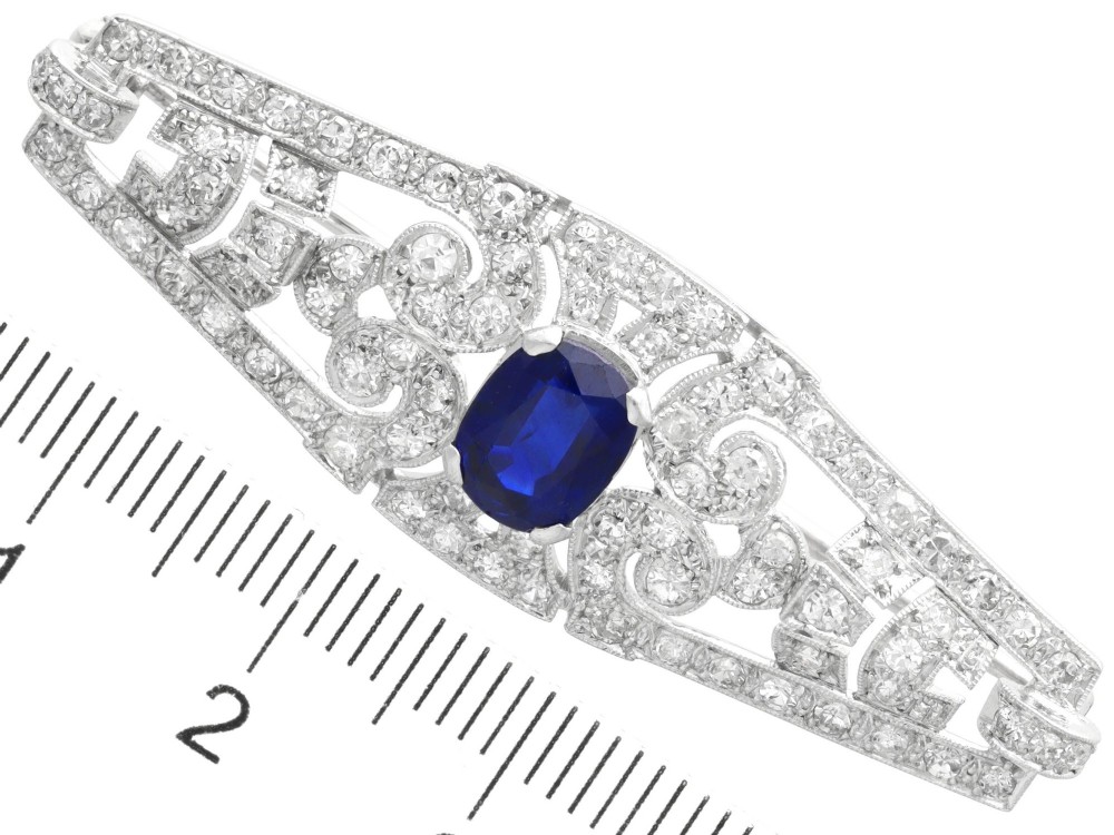 An Art Deco 9ct white gold brooch featuring a central oval blue sapphire framed by geometric openwork set with round diamonds. View IX