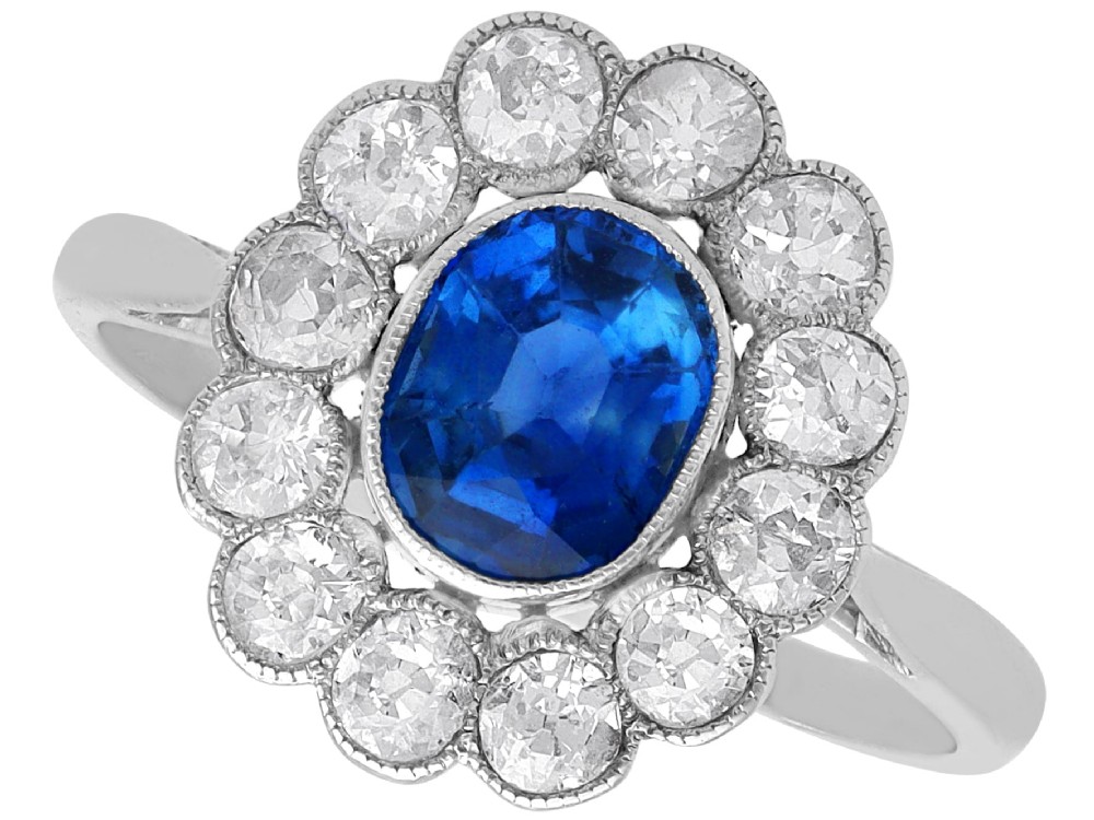 An antique platinum cluster ring featuring a central oval blue sapphire framed by twelve round cut diamonds with millegrain detail.