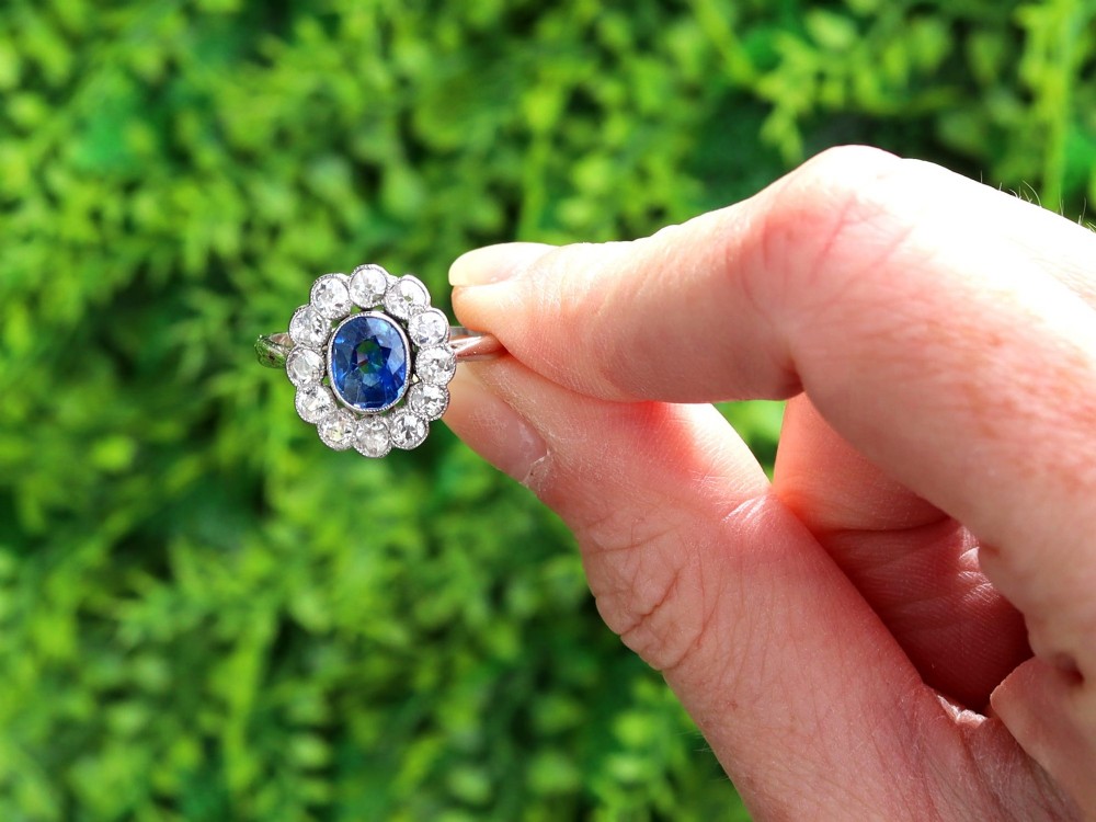 An antique platinum cluster ring featuring a central oval blue sapphire framed by twelve round cut diamonds with millegrain detail. View II