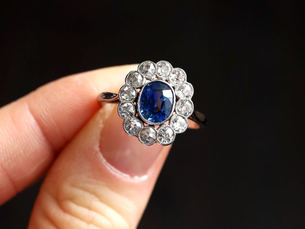 An antique platinum cluster ring featuring a central oval blue sapphire framed by twelve round cut diamonds with millegrain detail. View III