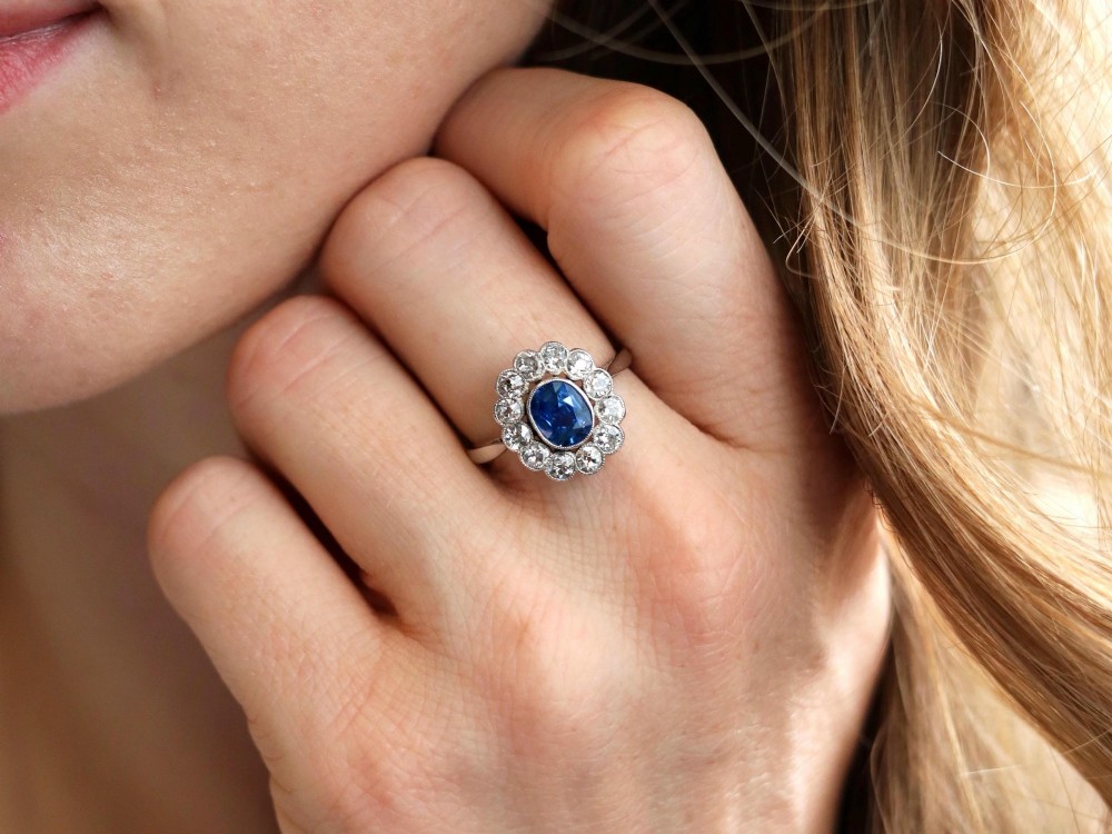 An antique platinum cluster ring featuring a central oval blue sapphire framed by twelve round cut diamonds with millegrain detail. View IV