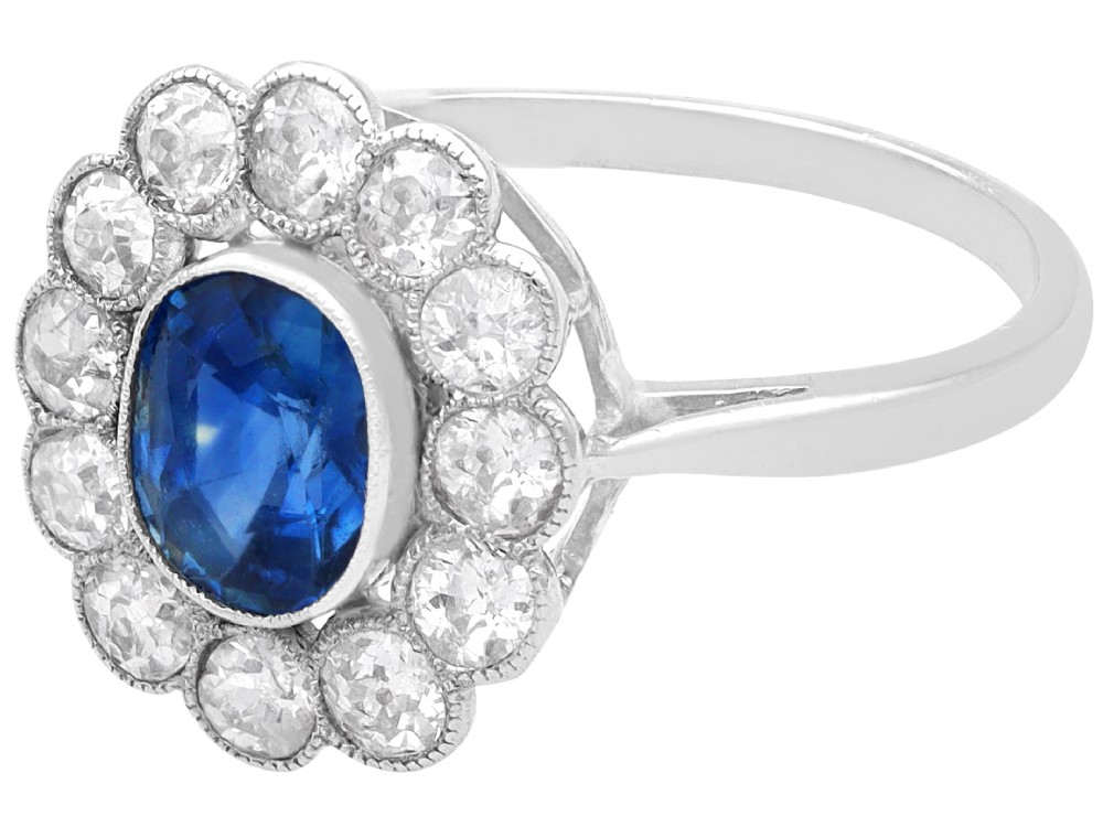 An antique platinum cluster ring featuring a central oval blue sapphire framed by twelve round cut diamonds with millegrain detail. View VI