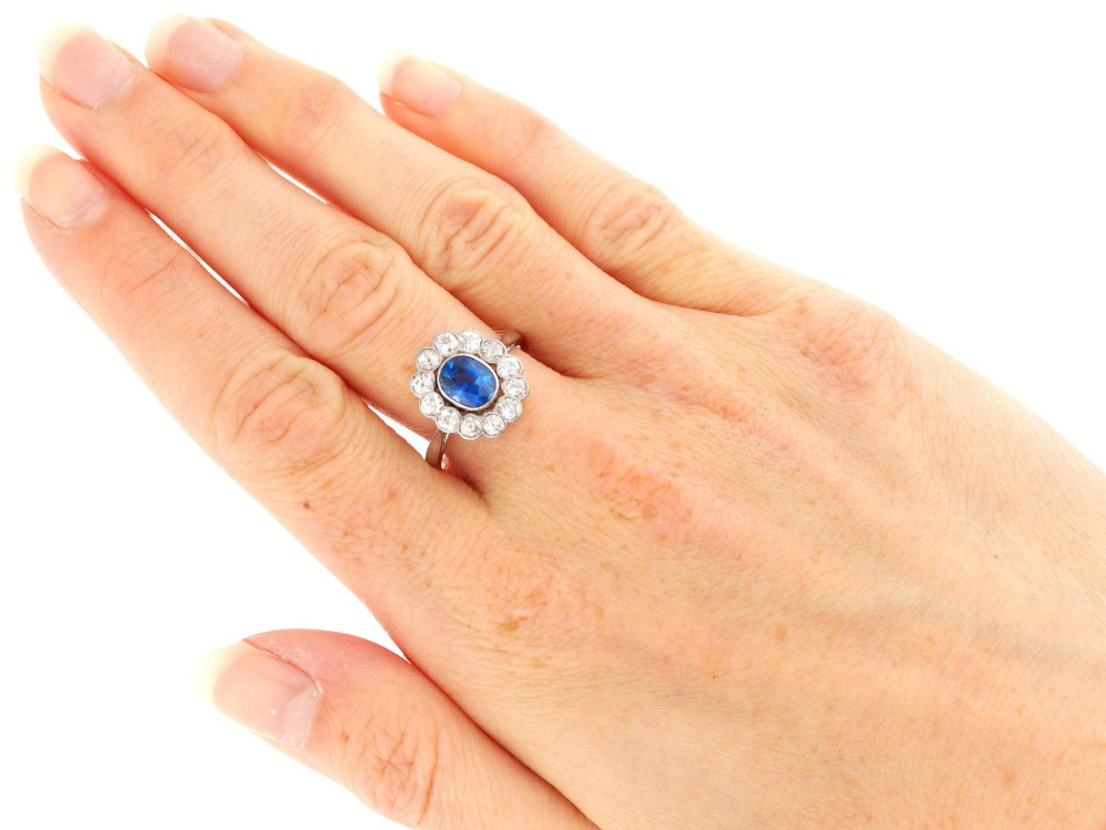 An antique platinum cluster ring featuring a central oval blue sapphire framed by twelve round cut diamonds with millegrain detail. View X