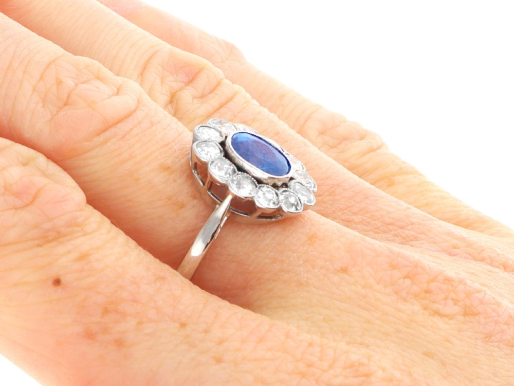 An antique platinum cluster ring featuring a central oval blue sapphire framed by twelve round cut diamonds with millegrain detail. View XI