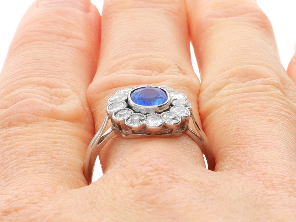 An antique platinum cluster ring featuring a central oval blue sapphire framed by twelve round cut diamonds with millegrain detail. View XII