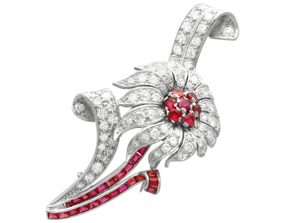 A platinum flower brooch featuring diamond-set petals, a cluster of rubies at the centre, and a stem lined with channel-set red rubies.