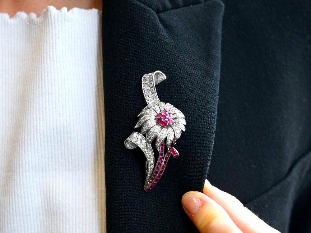 A platinum flower brooch featuring diamond-set petals, a cluster of rubies at the centre, and a stem lined with channel-set red rubies. View IV