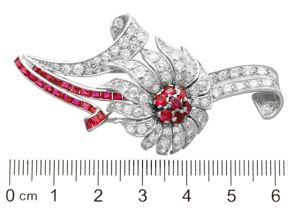 A platinum flower brooch featuring diamond-set petals, a cluster of rubies at the centre, and a stem lined with channel-set red rubies. View XI