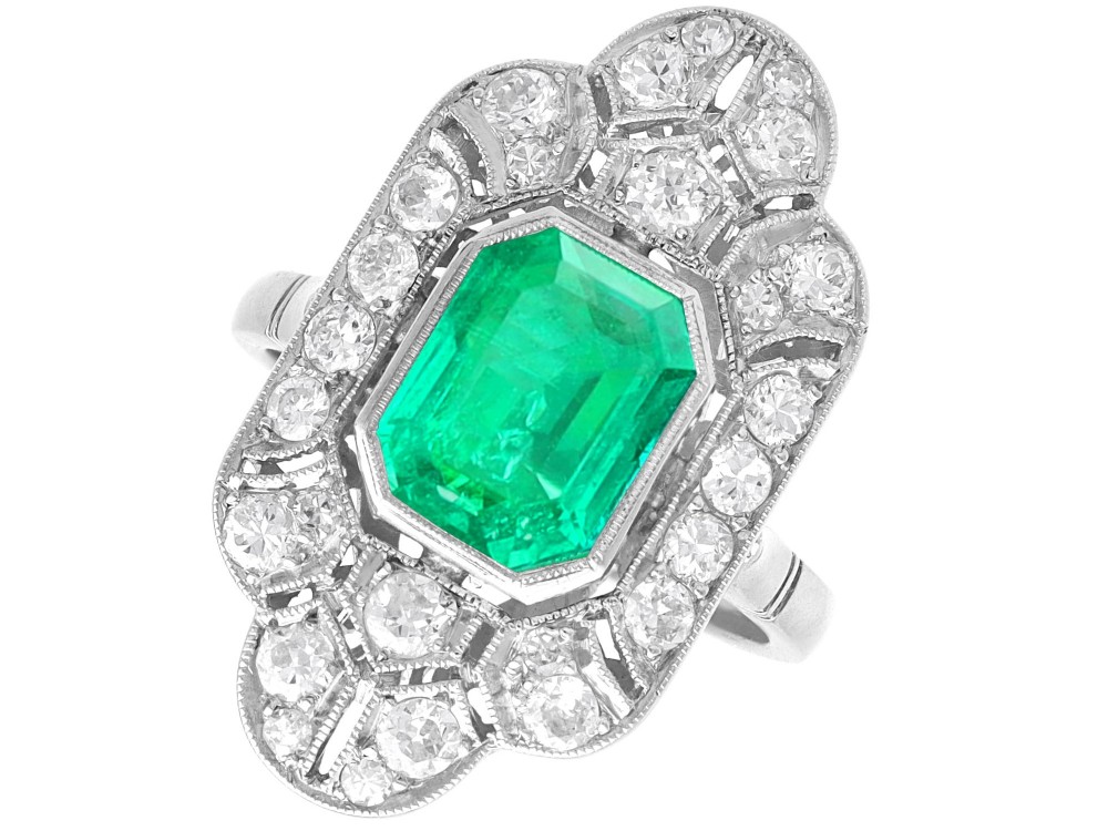 An Art Deco platinum dress ring featuring a central rectangular emerald surrounded by an elongated geometric pierced frame set with diamonds.