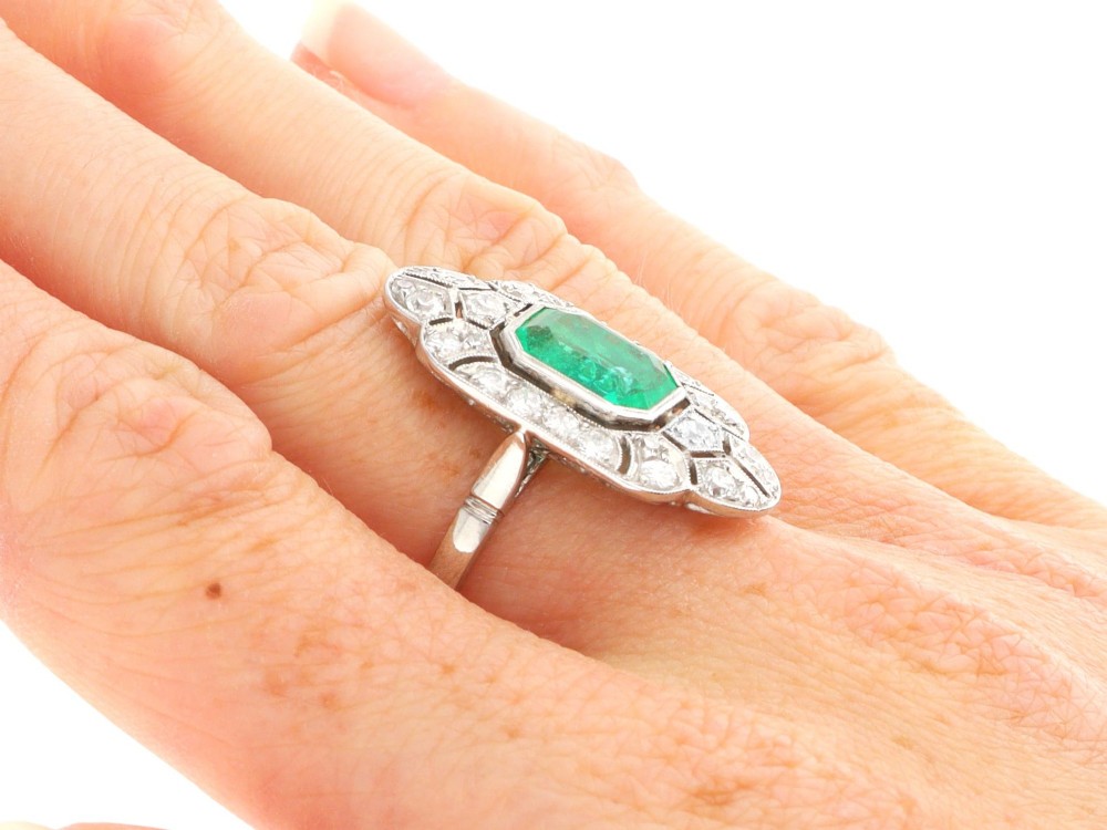 An Art Deco platinum dress ring featuring a central rectangular emerald surrounded by an elongated geometric pierced frame set with diamonds. View X