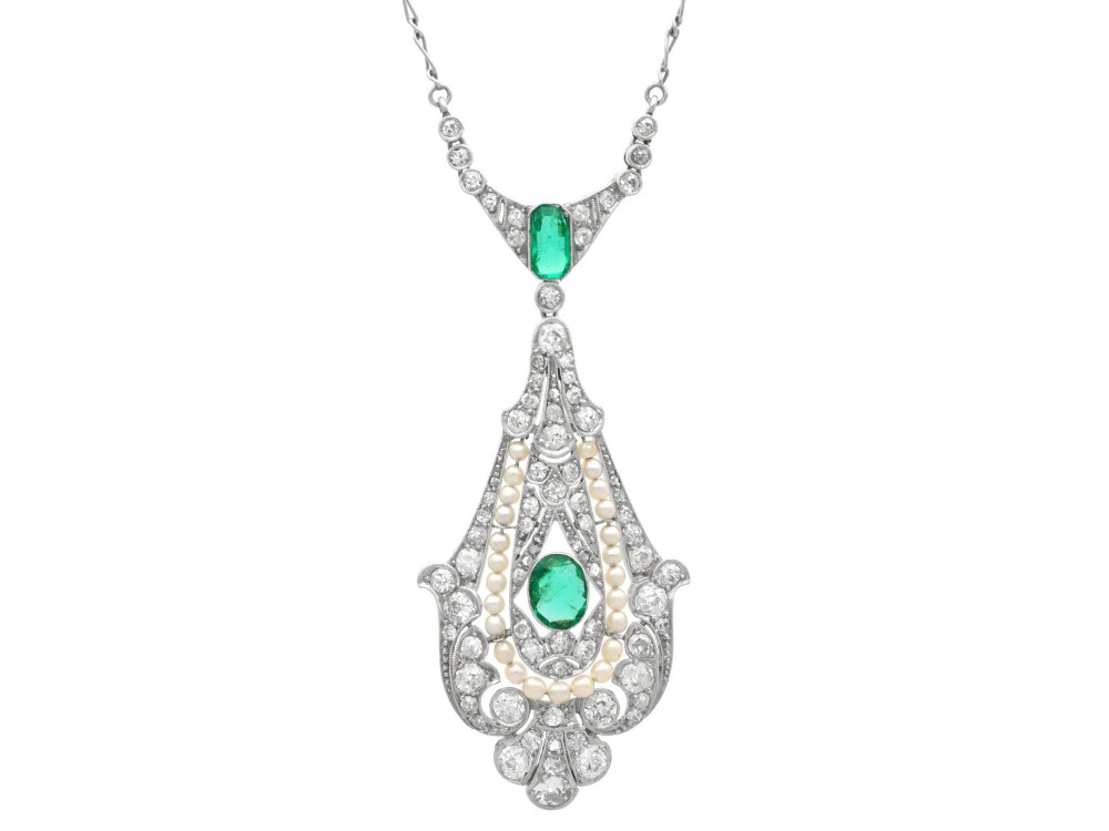An openwork platinum pendant featuring an elongated drop shape set with diamonds, a line of seed pearls, and two vibrant green Colombian emeralds.