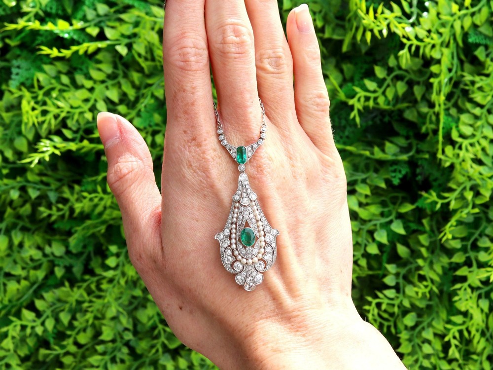 An openwork platinum pendant featuring an elongated drop shape set with diamonds, a line of seed pearls, and two vibrant green Colombian emeralds. View II