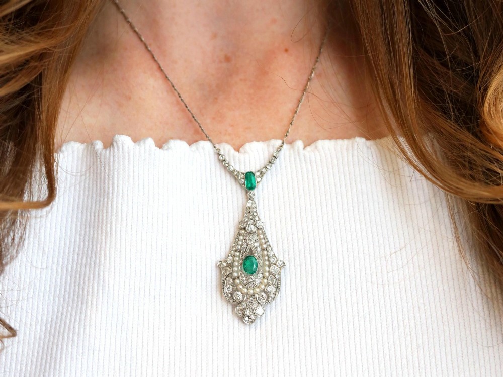 An openwork platinum pendant featuring an elongated drop shape set with diamonds, a line of seed pearls, and two vibrant green Colombian emeralds. View IV