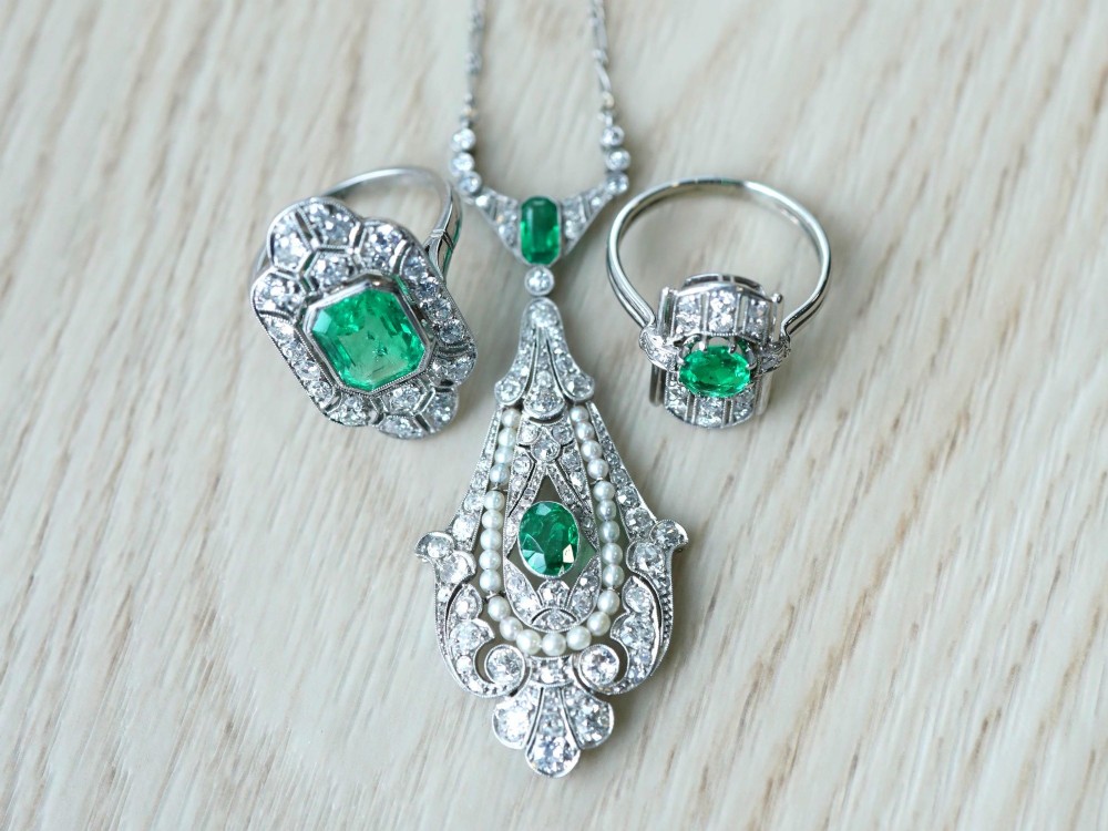 An openwork platinum pendant featuring an elongated drop shape set with diamonds, a line of seed pearls, and two vibrant green Colombian emeralds. View VI