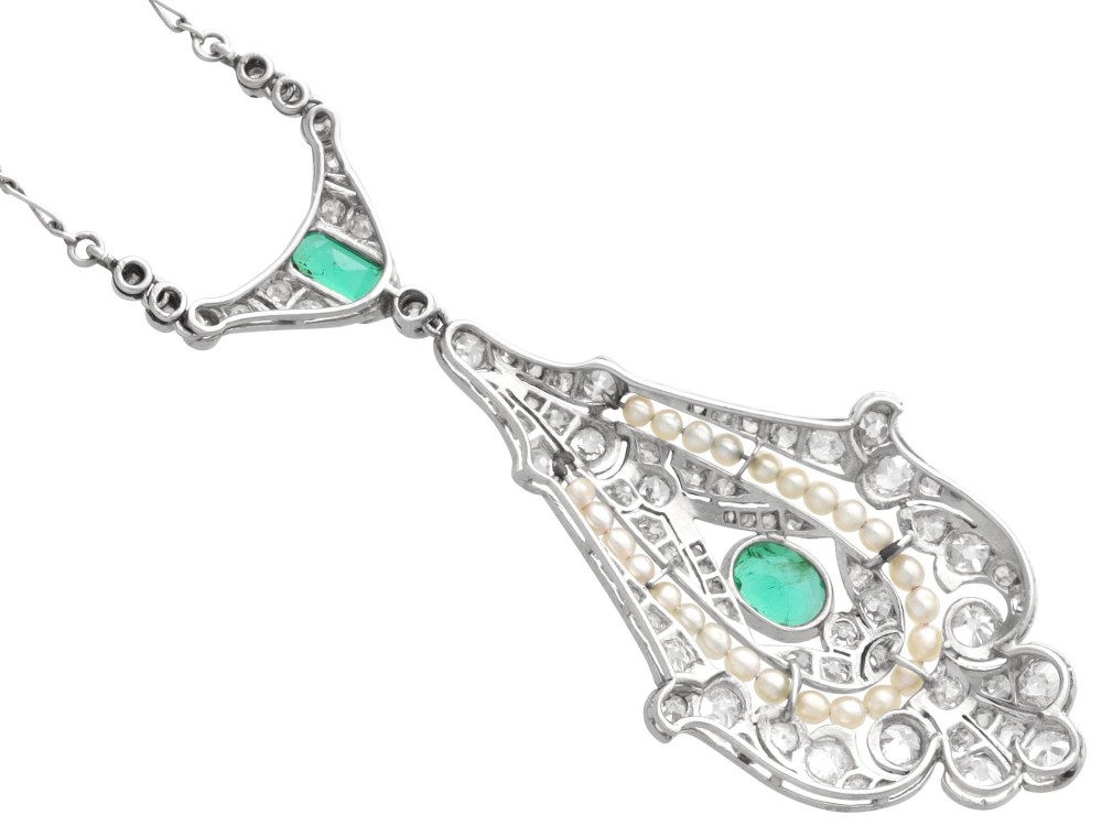 An openwork platinum pendant featuring an elongated drop shape set with diamonds, a line of seed pearls, and two vibrant green Colombian emeralds. View X