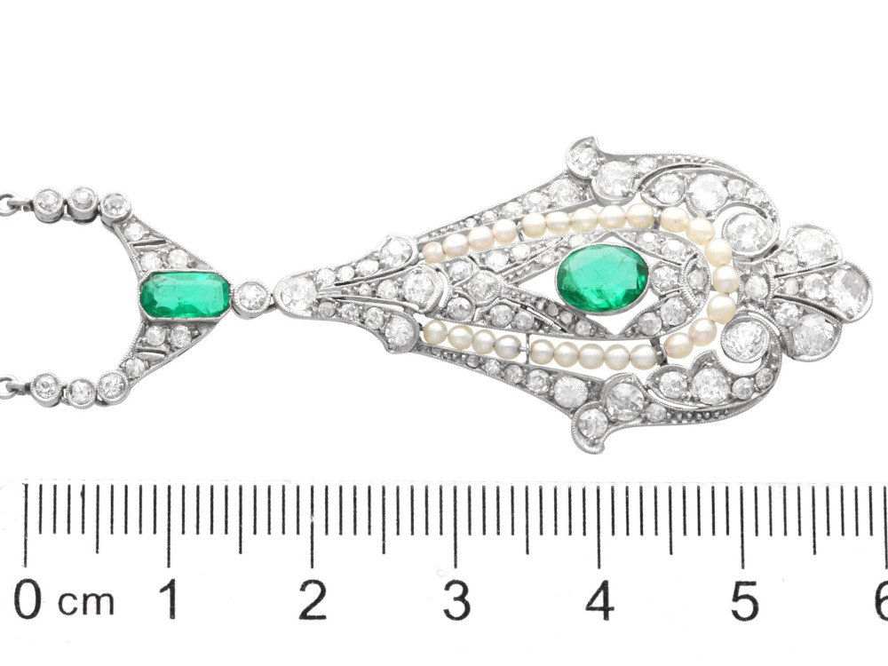 An openwork platinum pendant featuring an elongated drop shape set with diamonds, a line of seed pearls, and two vibrant green Colombian emeralds. View XI