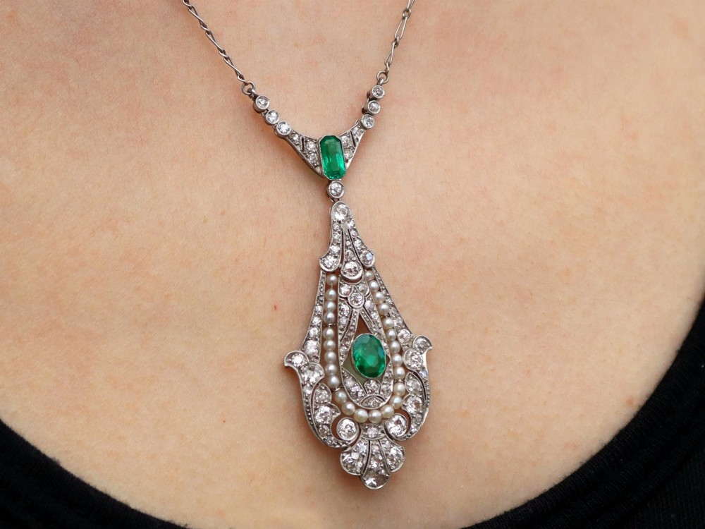 An openwork platinum pendant featuring an elongated drop shape set with diamonds, a line of seed pearls, and two vibrant green Colombian emeralds. View XII