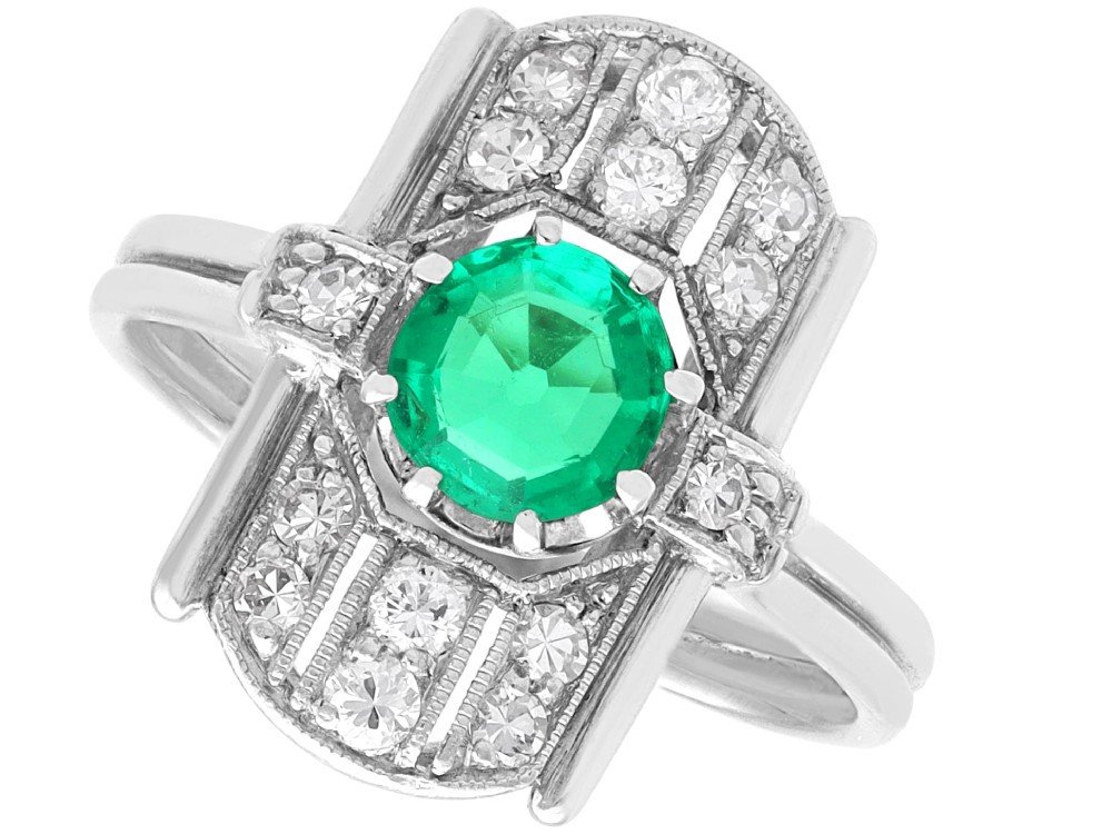 An Art Deco 14ct white gold ring featuring a central round green emerald framed by geometric shoulders set with small brilliant diamonds.