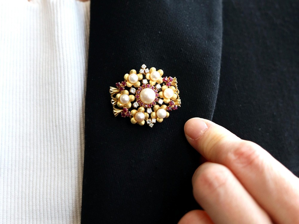 An 18ct yellow gold floral cluster brooch set with a central pearl surrounded by small rubies, outer cultured pearls, round diamonds, and red rubies. View IV