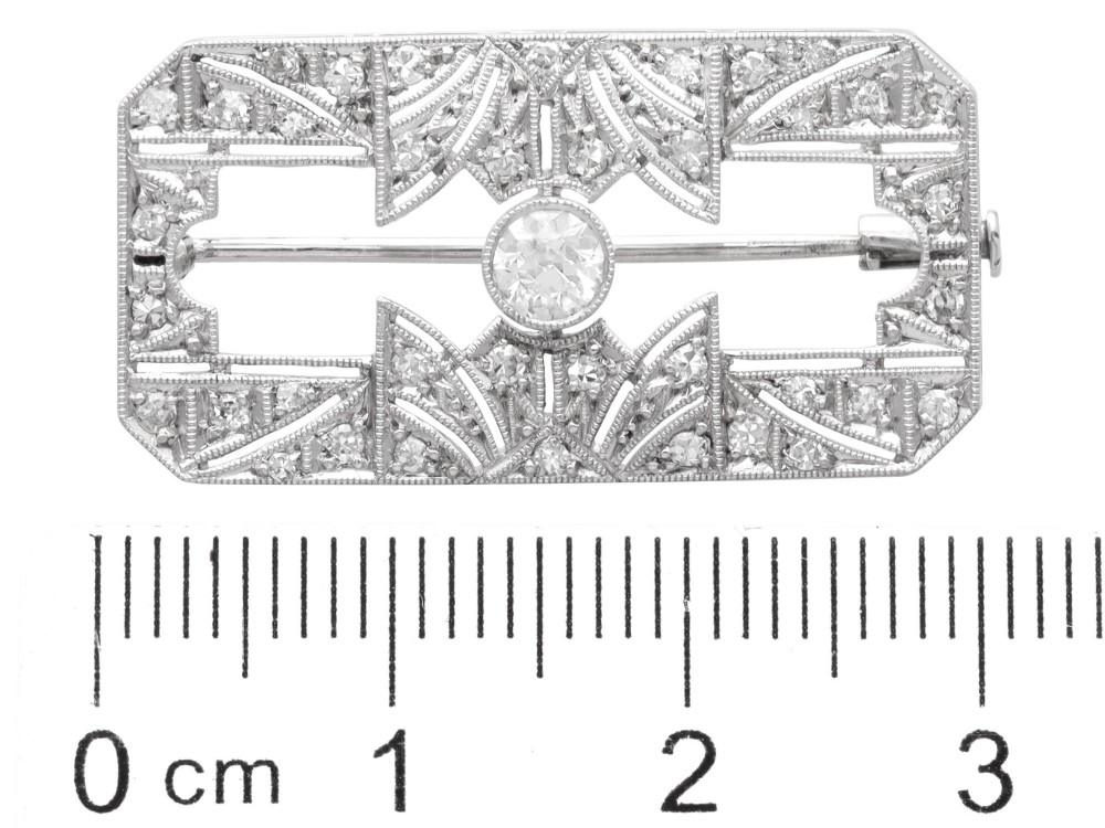A rectangular Art Deco 18ct white gold brooch featuring openwork geometric filigree, millegrain edging, and a central round-cut diamond. View XI