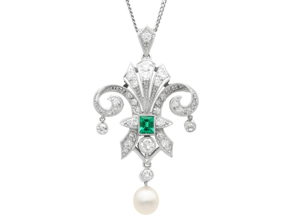 An Art Deco platinum pendant shaped as a stylised fleur-de-lis, embellished with pave-set diamonds, a central rectangular emerald, and a hanging pearl drop.