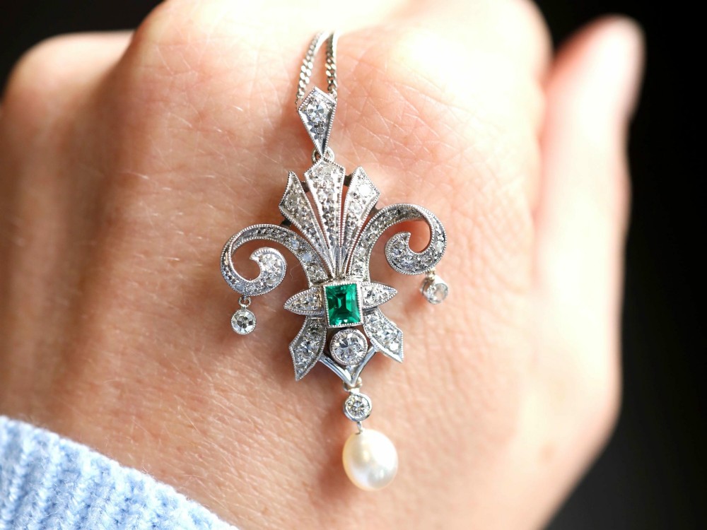 An Art Deco platinum pendant shaped as a stylised fleur-de-lis, embellished with pave-set diamonds, a central rectangular emerald, and a hanging pearl drop. View III