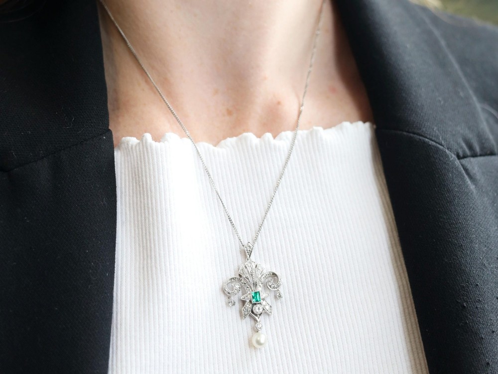 An Art Deco platinum pendant shaped as a stylised fleur-de-lis, embellished with pave-set diamonds, a central rectangular emerald, and a hanging pearl drop. View IV