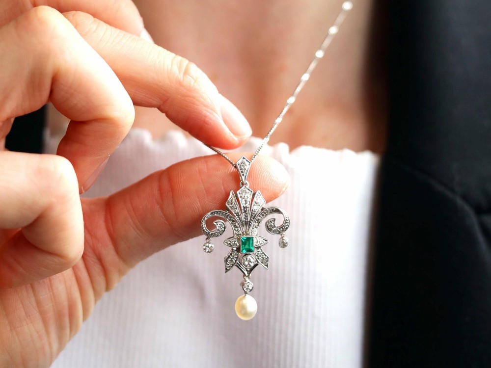 An Art Deco platinum pendant shaped as a stylised fleur-de-lis, embellished with pave-set diamonds, a central rectangular emerald, and a hanging pearl drop. View V