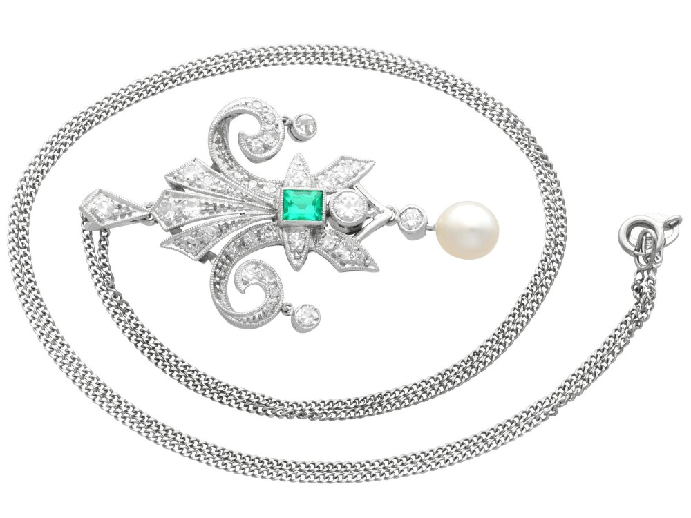 An Art Deco platinum pendant shaped as a stylised fleur-de-lis, embellished with pave-set diamonds, a central rectangular emerald, and a hanging pearl drop. View VII