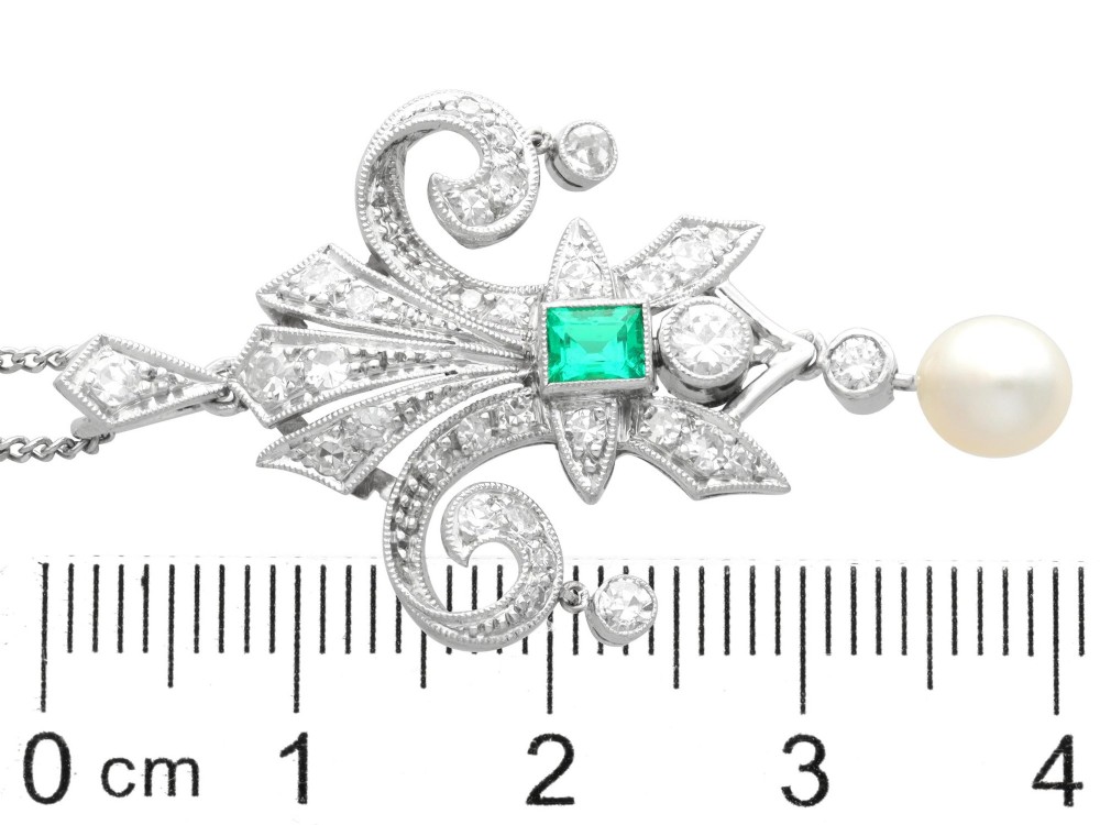 An Art Deco platinum pendant shaped as a stylised fleur-de-lis, embellished with pave-set diamonds, a central rectangular emerald, and a hanging pearl drop. View X