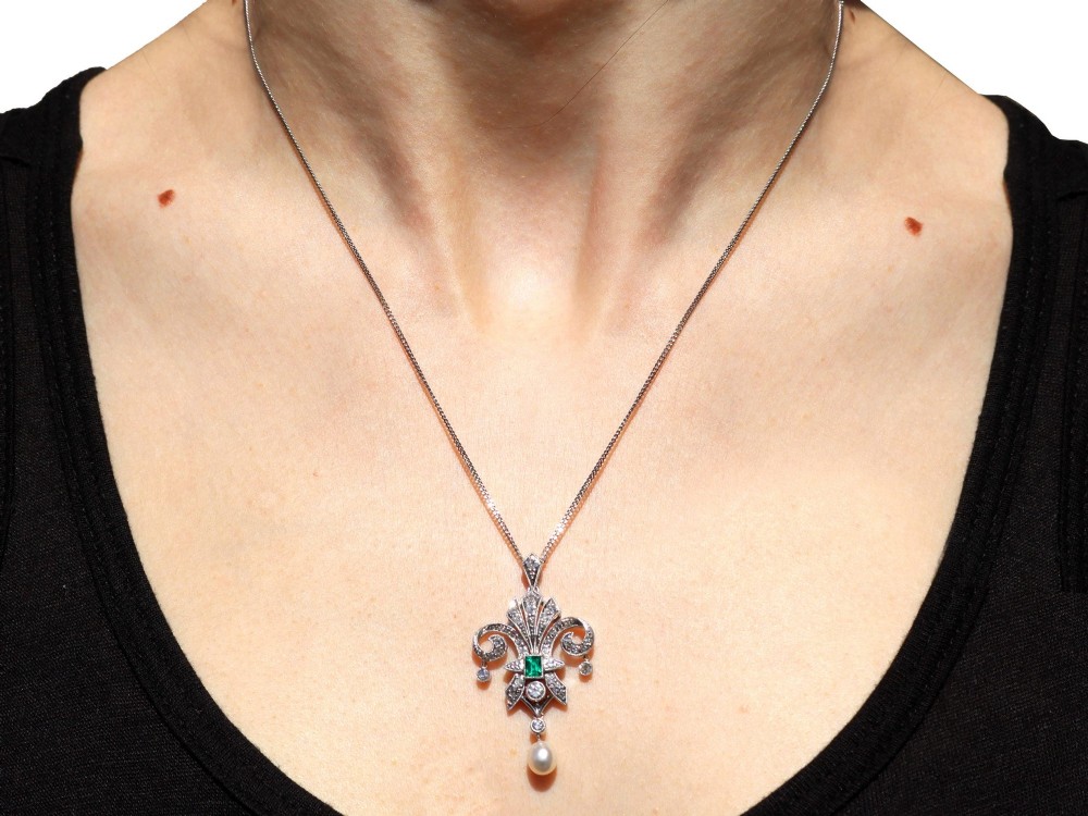 An Art Deco platinum pendant shaped as a stylised fleur-de-lis, embellished with pave-set diamonds, a central rectangular emerald, and a hanging pearl drop. View XII