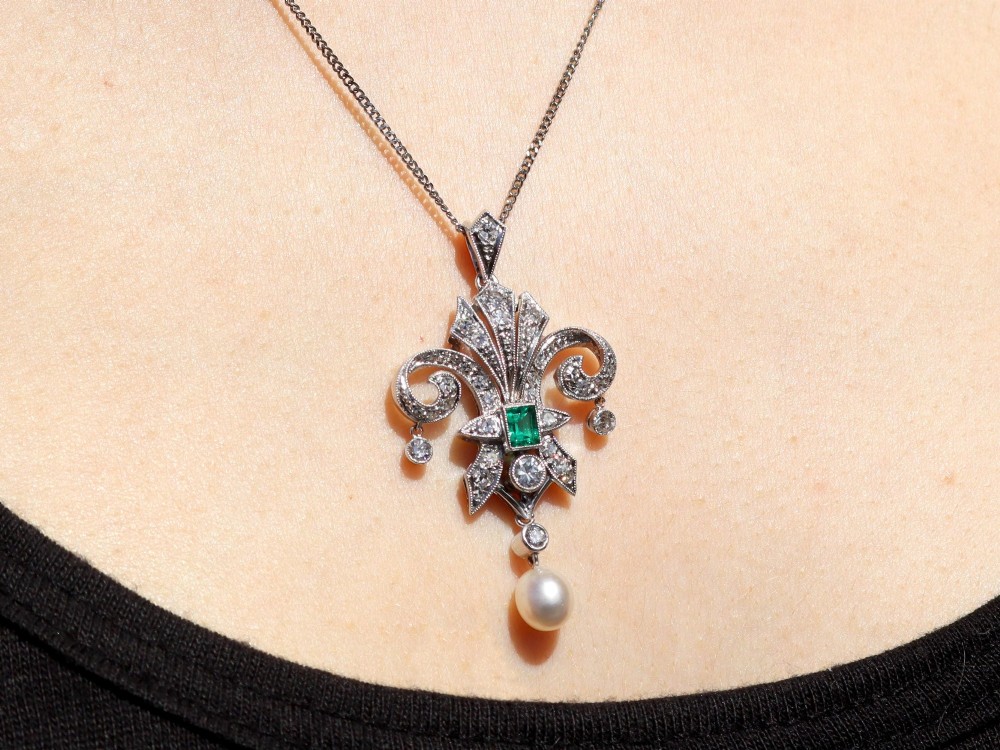 An Art Deco platinum pendant shaped as a stylised fleur-de-lis, embellished with pave-set diamonds, a central rectangular emerald, and a hanging pearl drop. View XIII