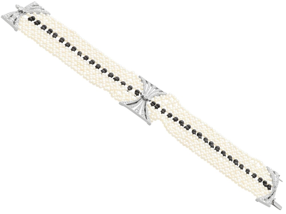 An Art Deco multi-strand pearl and black onyx cuff bracelet featuring a central geometric bow-shaped platinum mount set with diamonds. View X