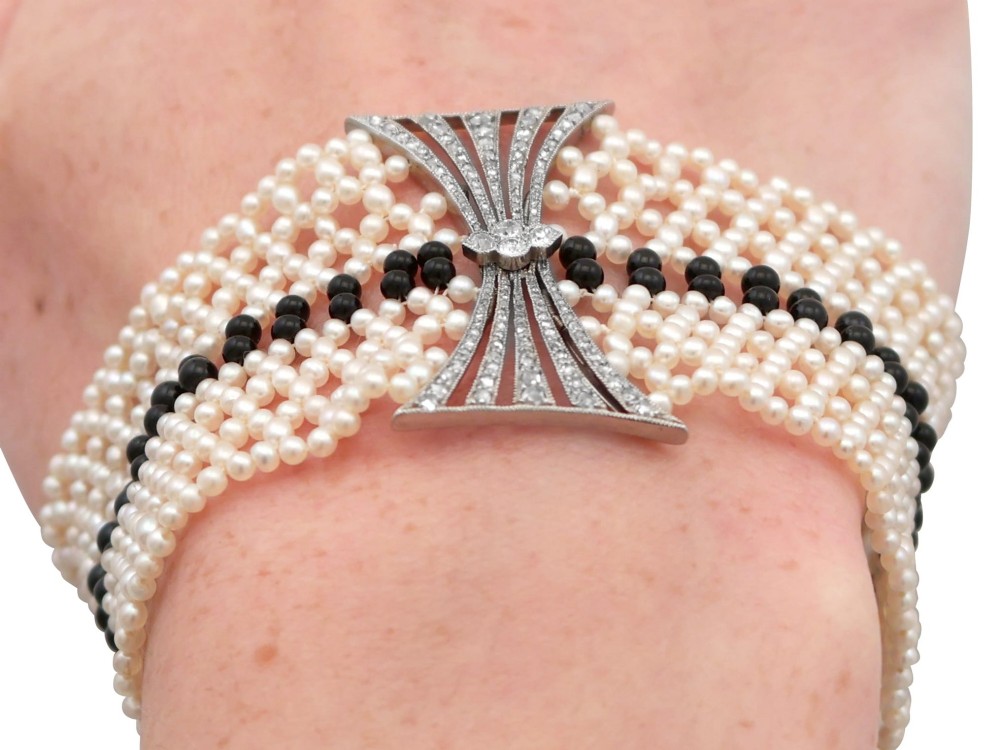 An Art Deco multi-strand pearl and black onyx cuff bracelet featuring a central geometric bow-shaped platinum mount set with diamonds. View XV