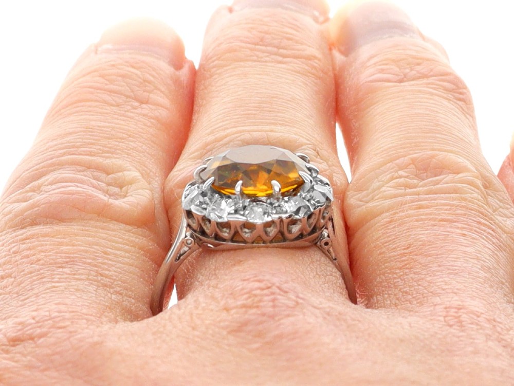 An antique Art Deco platinum ring featuring a central round orange zircon surrounded by a halo of claw-set diamonds. View XIV