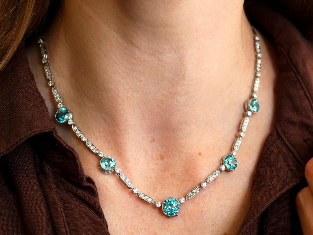An Art Deco platinum chain necklace featuring five bezel-set round blue zircons separated by geometric diamond-set bar links. View VI