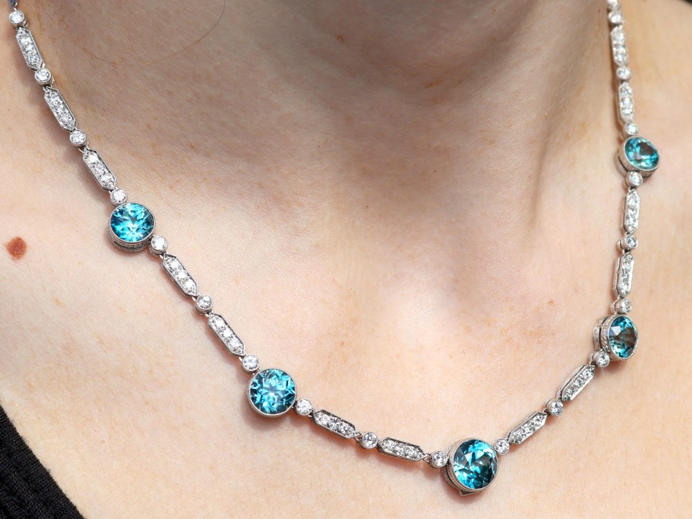 An Art Deco platinum chain necklace featuring five bezel-set round blue zircons separated by geometric diamond-set bar links. View XIV