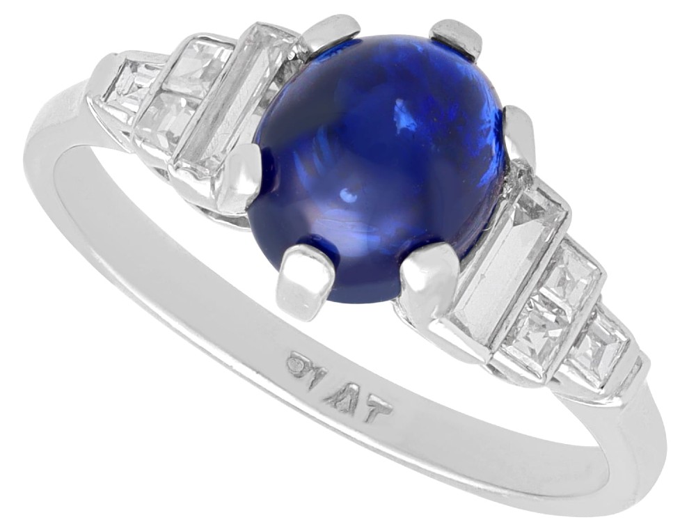 An Art Deco platinum dress ring set with a central oval cabochon blue sapphire flanked by stepped geometric shoulders of baguette cut diamonds.