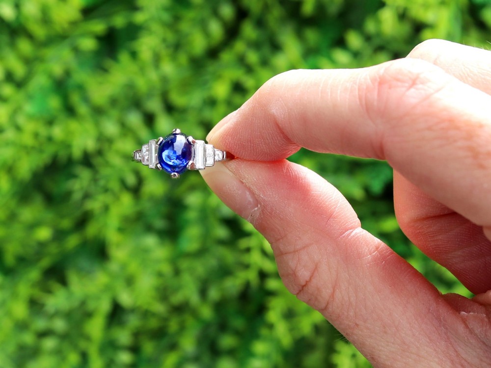 An Art Deco platinum dress ring set with a central oval cabochon blue sapphire flanked by stepped geometric shoulders of baguette cut diamonds. View II