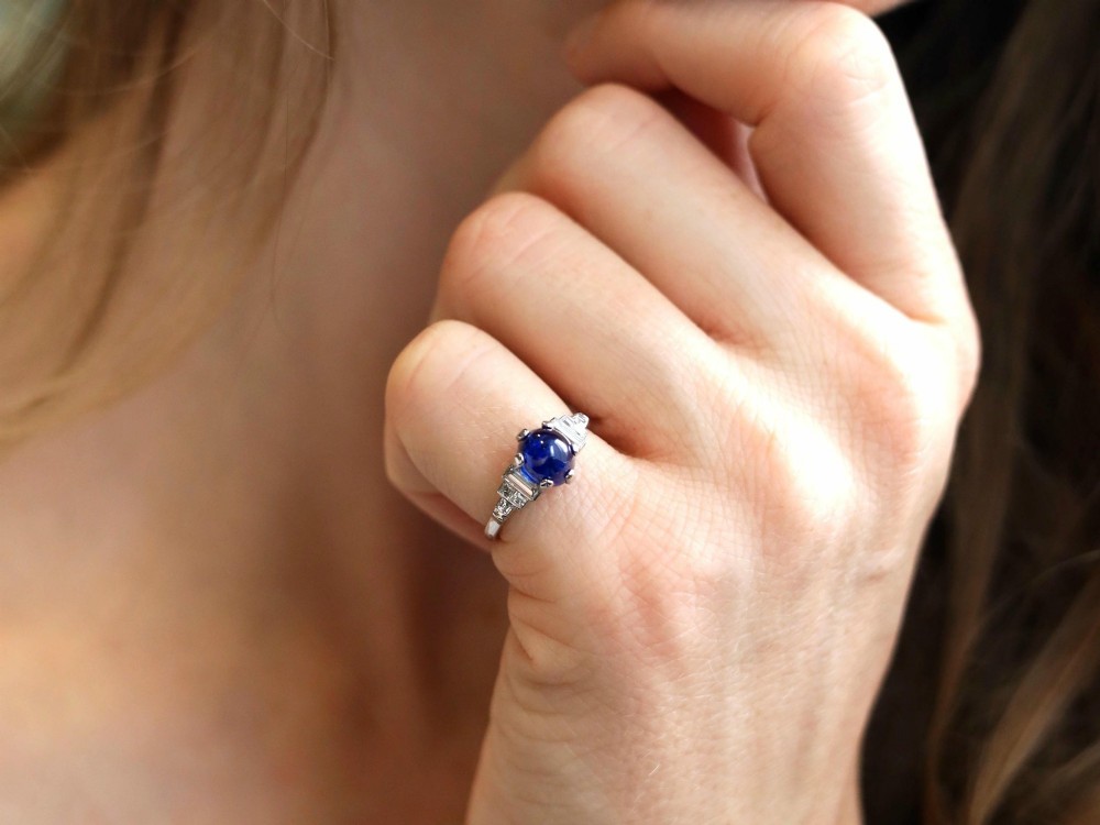 An Art Deco platinum dress ring set with a central oval cabochon blue sapphire flanked by stepped geometric shoulders of baguette cut diamonds. View III