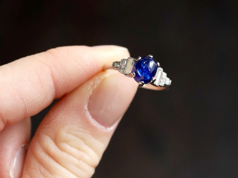 An Art Deco platinum dress ring set with a central oval cabochon blue sapphire flanked by stepped geometric shoulders of baguette cut diamonds. View IV