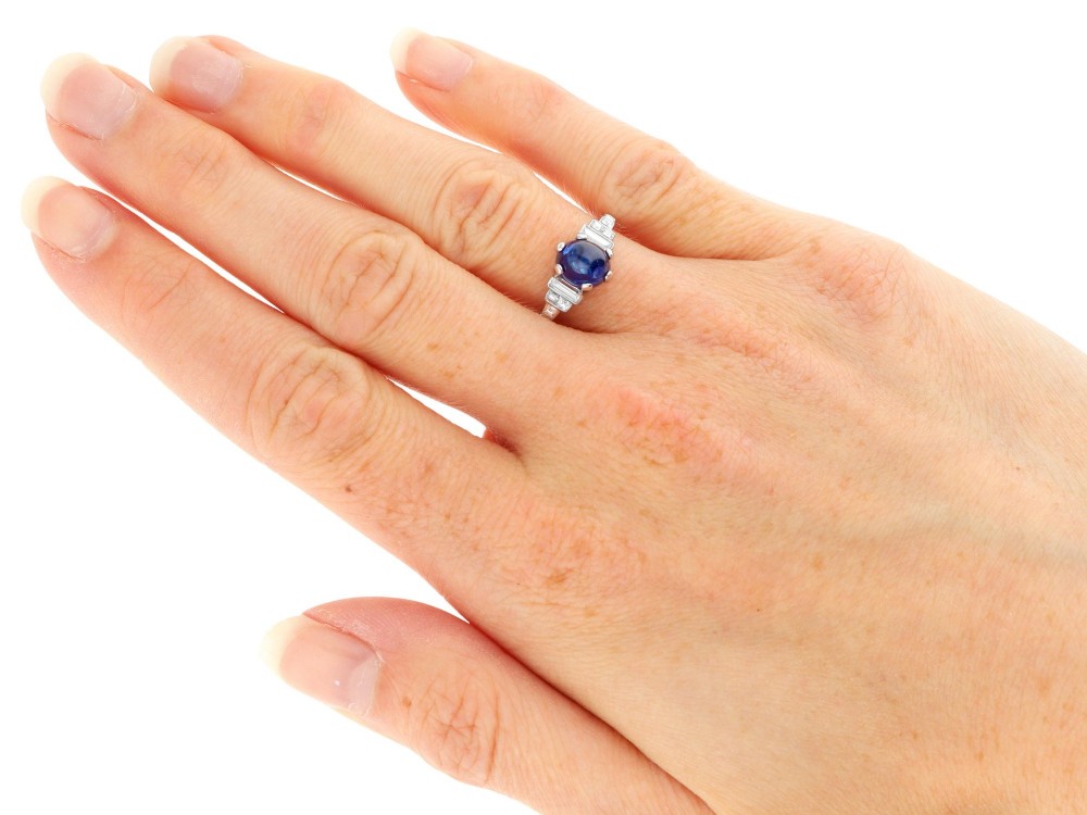 An Art Deco platinum dress ring set with a central oval cabochon blue sapphire flanked by stepped geometric shoulders of baguette cut diamonds. View X